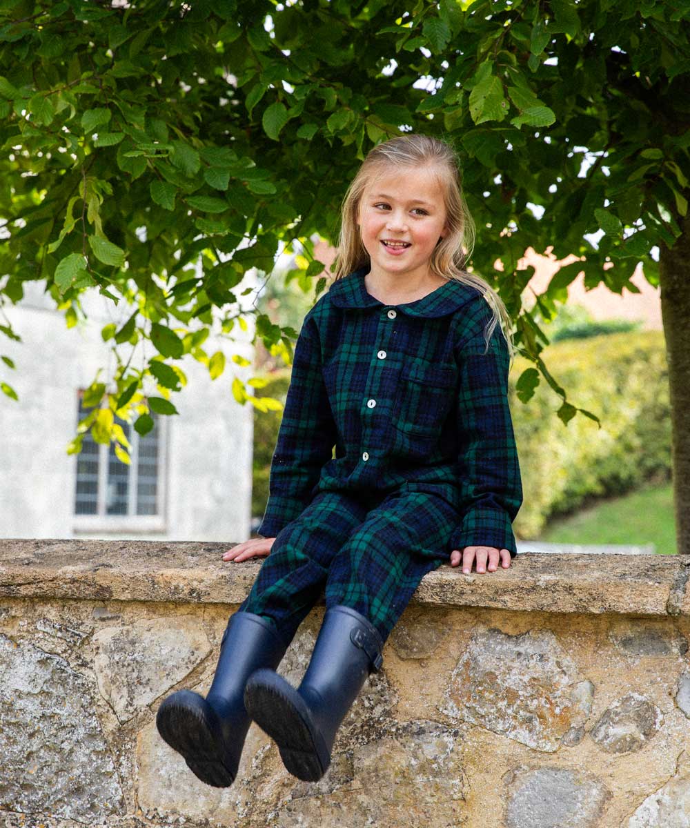Young girl in black watch tartan pyjamas and rain boots sitting on a stone wall with greenery in the background