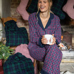 Woman in plaid pajamas holding a mug in a festive setting with stockings and a fireplace.