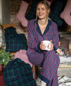 Woman in plaid pajamas holding a mug in a festive setting with stockings and a fireplace.
