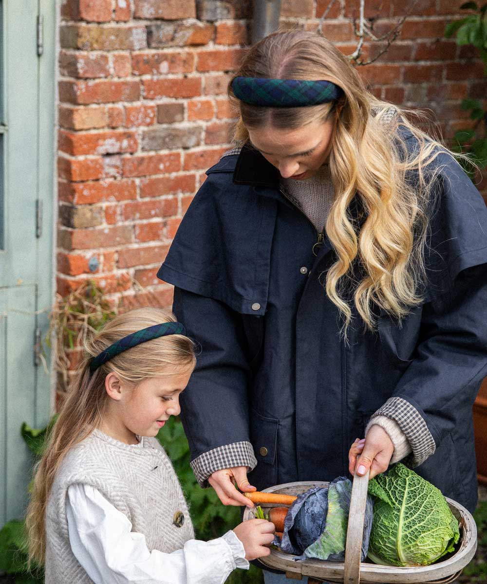 Woman and child outdoors with a basket of vegetables against a brick wall.