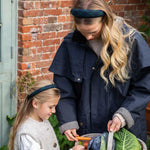 Woman and child outdoors with a basket of vegetables against a brick wall.