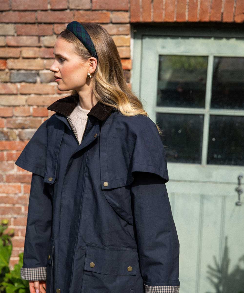 Woman wearing a navy blue coat standing in front of a brick wall and glass door wearing a tartan headband