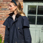 Woman wearing a navy blue coat standing in front of a brick wall and glass door wearing a tartan headband