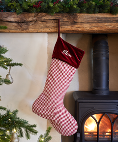 Red checkered Christmas stocking with velvet top hanging by a fireplace.