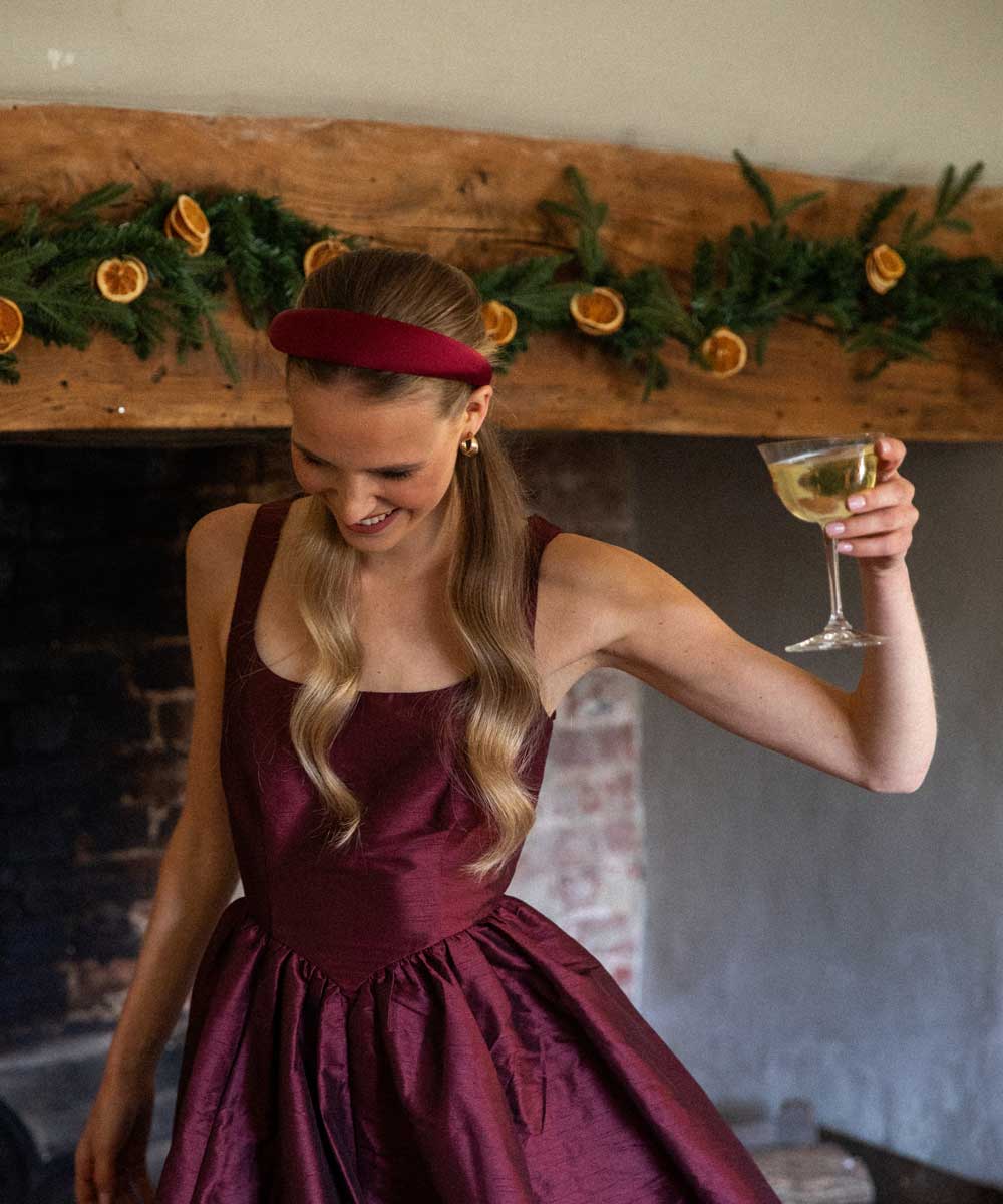 Woman in a burgundy dress and burgundy satin headband holding a glass of white wine in front of a decorated fireplace.