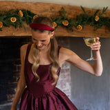 Woman in a burgundy dress and burgundy satin headband holding a glass of white wine in front of a decorated fireplace.