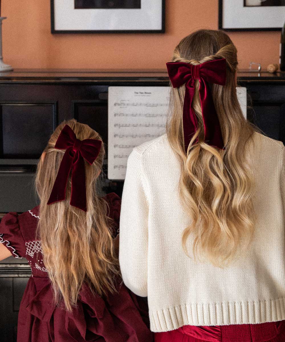Two people with long blonde hair wearing burgundy velvet bows, standing in front of a piano with sheet music.