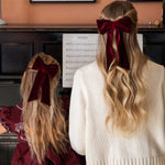 Two people with long blonde hair wearing burgundy velvet bows, standing in front of a piano with sheet music.