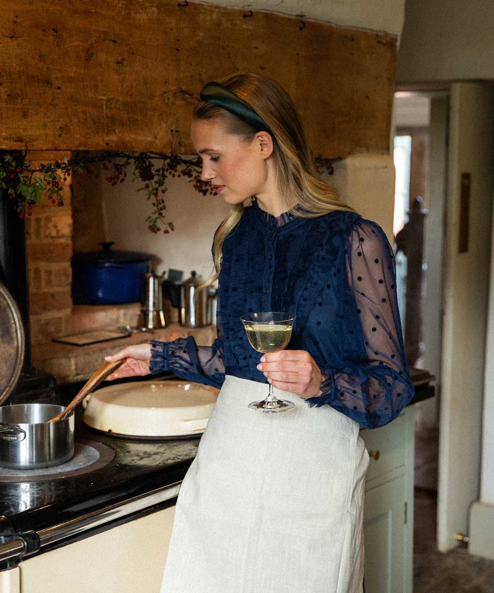 Woman in a kitchen holding a glass of wine, wearing a green satin headband and navy blue top and white apron.