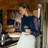 Woman in a kitchen holding a glass of wine, wearing a green satin headband and navy blue top and white apron.