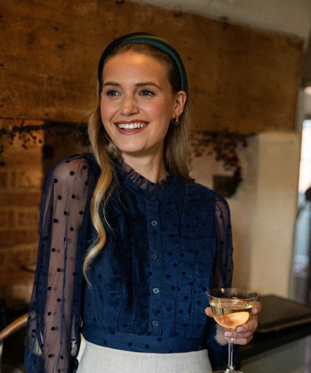 Woman in a dark blue blouse with a dark greens satin headband holding a cocktail in a warm-toned indoor setting.