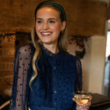 Woman in a dark blue blouse with a dark greens satin headband holding a cocktail in a warm-toned indoor setting.