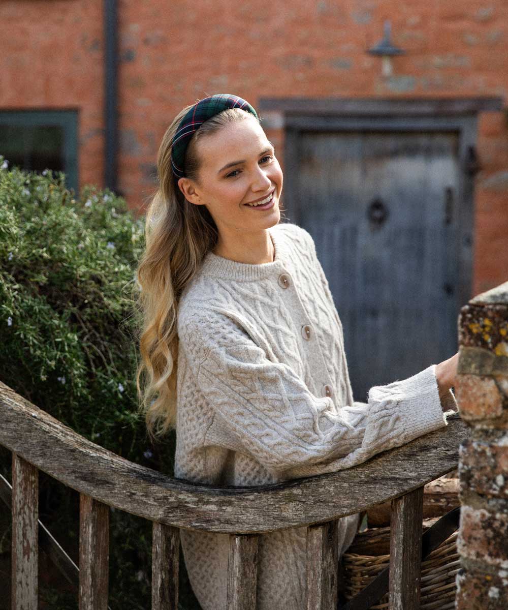 Woman wearing a tartan headband and cream cable knit sweater sitting on a wooden bench outdoors.