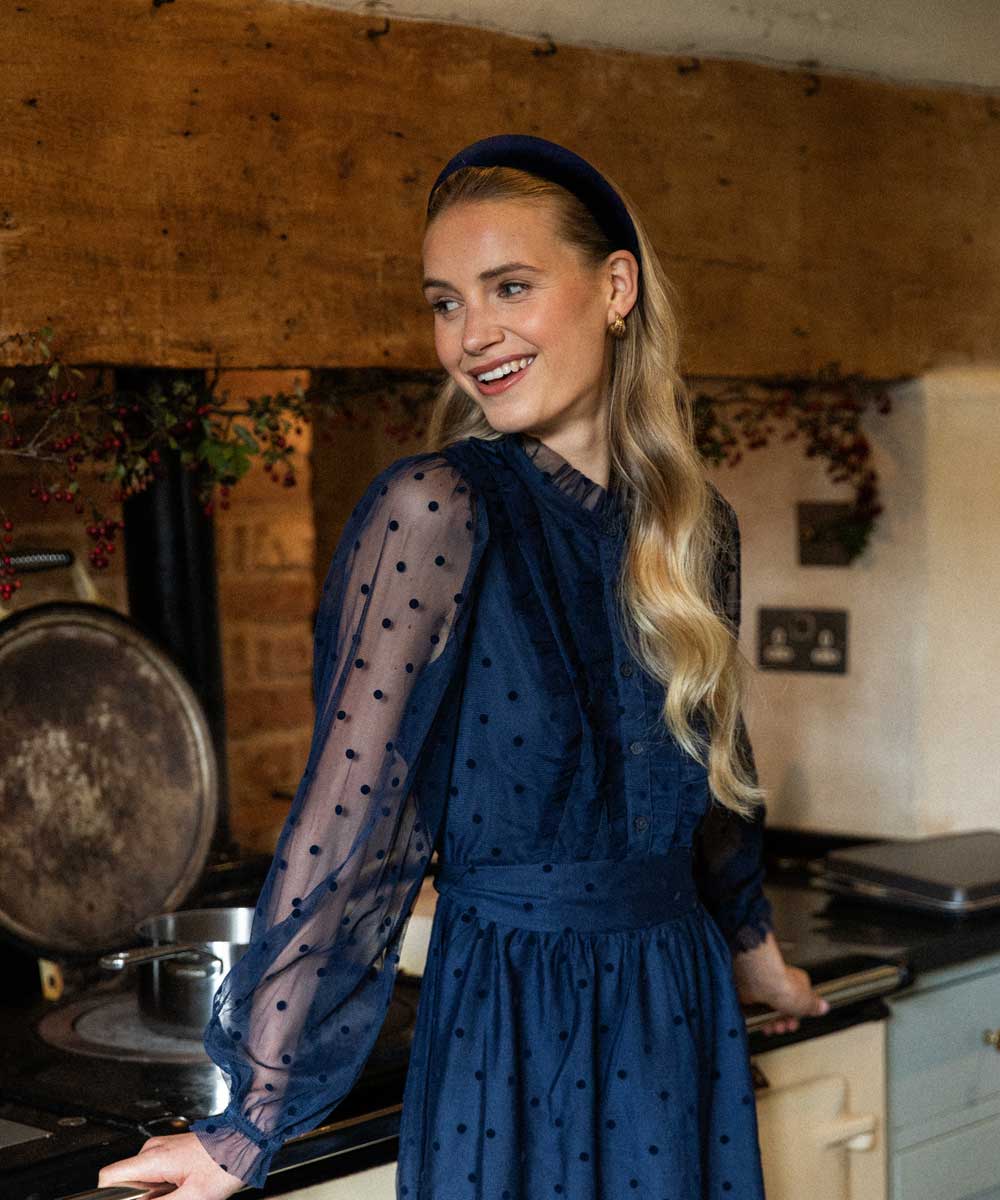 Woman in a blue dress and blue velvet headband standing in a rustic kitchen.