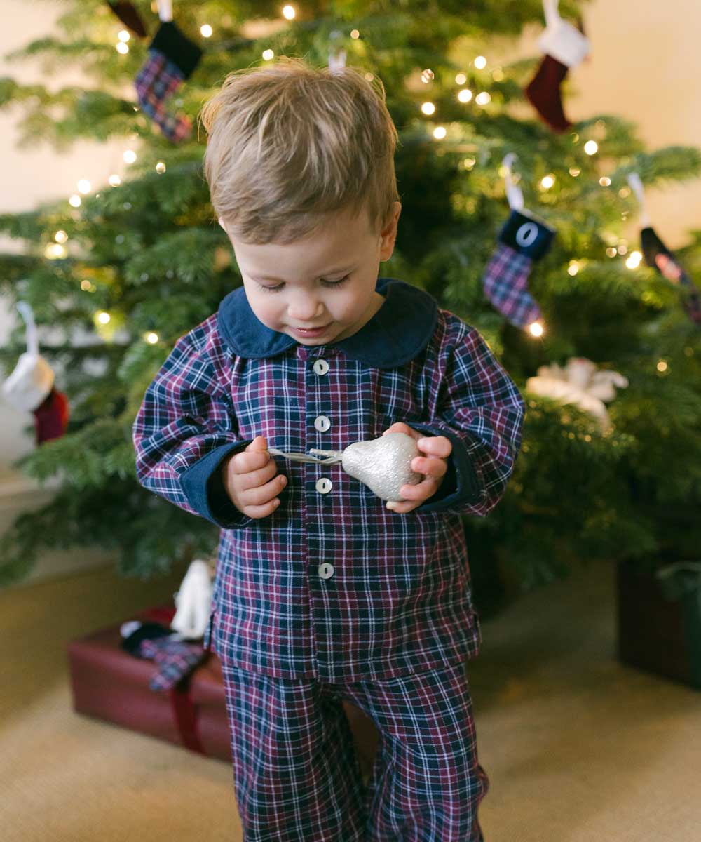 A child in a Christmas-themed brushed cotton tartan pyjama set with a contrasting Peter Pan collar, standing in front of a Christmas tree.