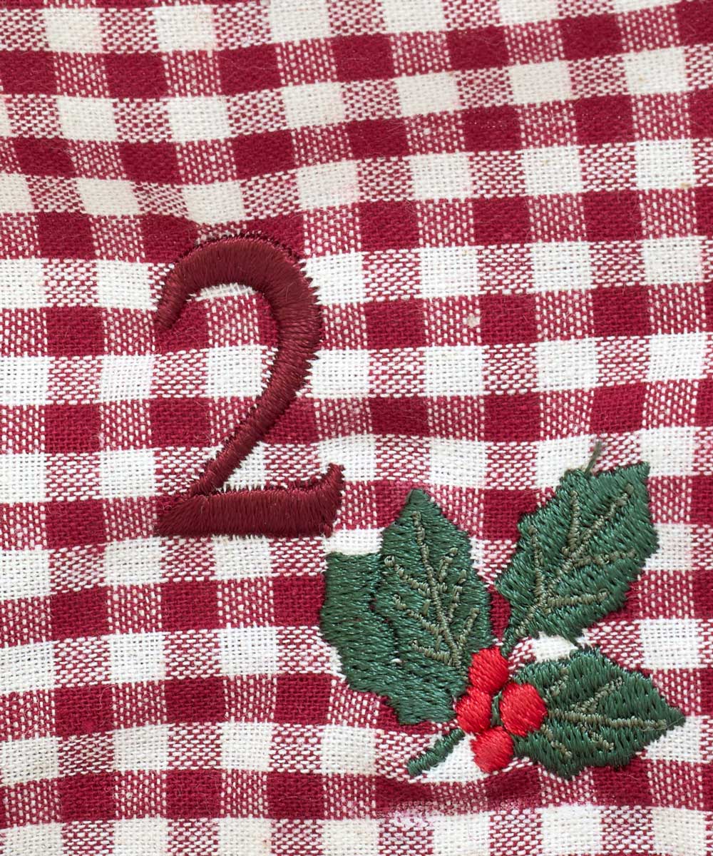A close-up of a red and white gingham fabric with embroidered details, featuring a dark red embroidered number '2' and a holly leaf and berry motif.