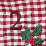 A close-up of a red and white gingham fabric with embroidered details, featuring a dark red embroidered number '2' and a holly leaf and berry motif.