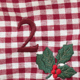 A close-up of a red and white gingham fabric with embroidered details, featuring a dark red embroidered number '2' and a holly leaf and berry motif.