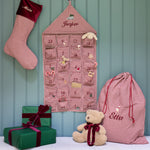 A festive Christmas advent setup featuring a pink and white gingham stocking, a matching house-shaped advent calendar, a large drawstring gift bag, and wrapped presents on a bench with a teddy bear, all set against a teal paneled wall.