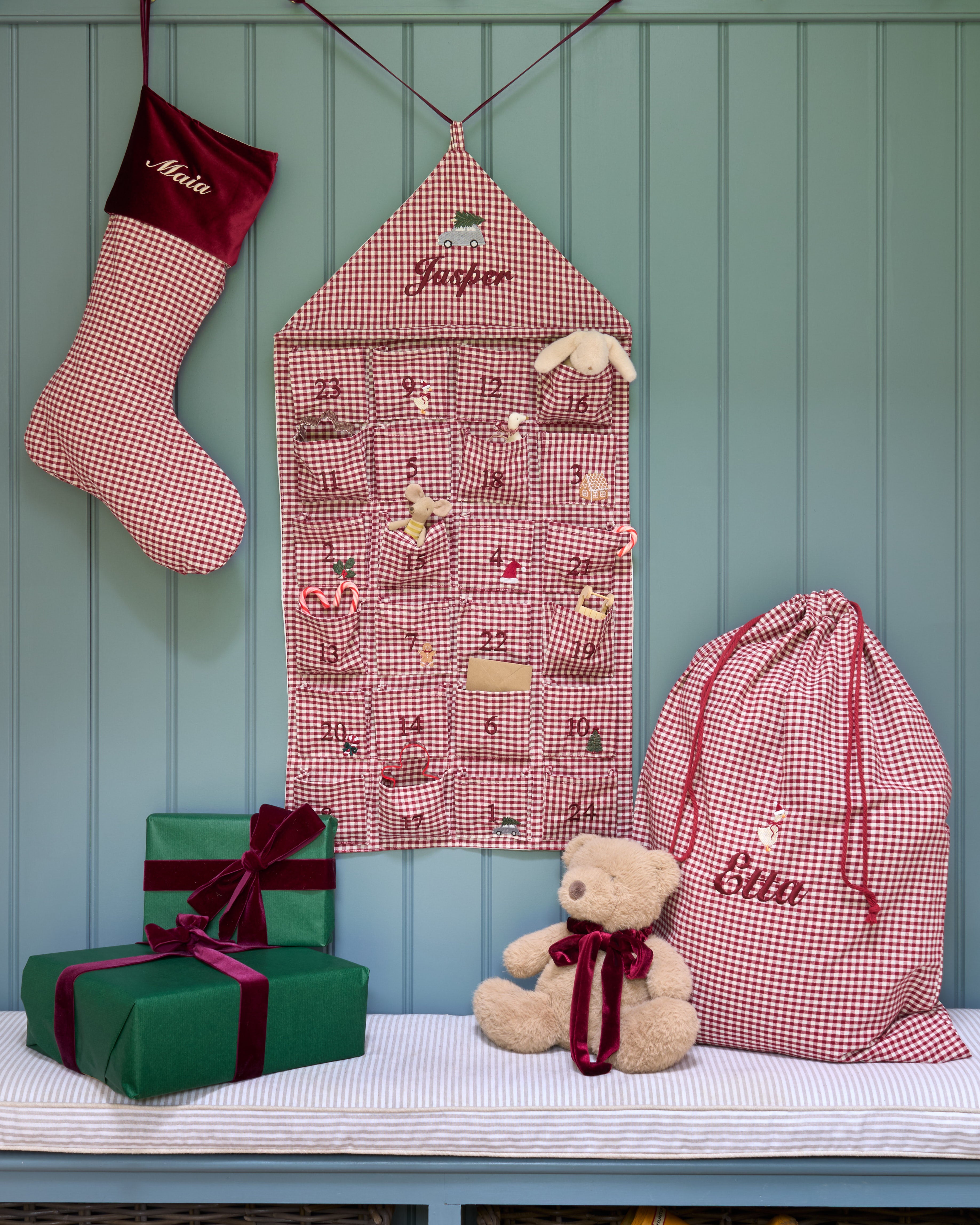 A festive Christmas advent setup featuring a pink and white gingham stocking, a matching house-shaped advent calendar, a large drawstring gift bag, and wrapped presents on a bench with a teddy bear, all set against a teal paneled wall.