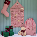A festive Christmas advent setup featuring a burgundy and white gingham stocking, a matching house-shaped advent calendar, a large drawstring gift bag, and wrapped presents on a bench with a teddy bear, all set against a teal paneled wall.