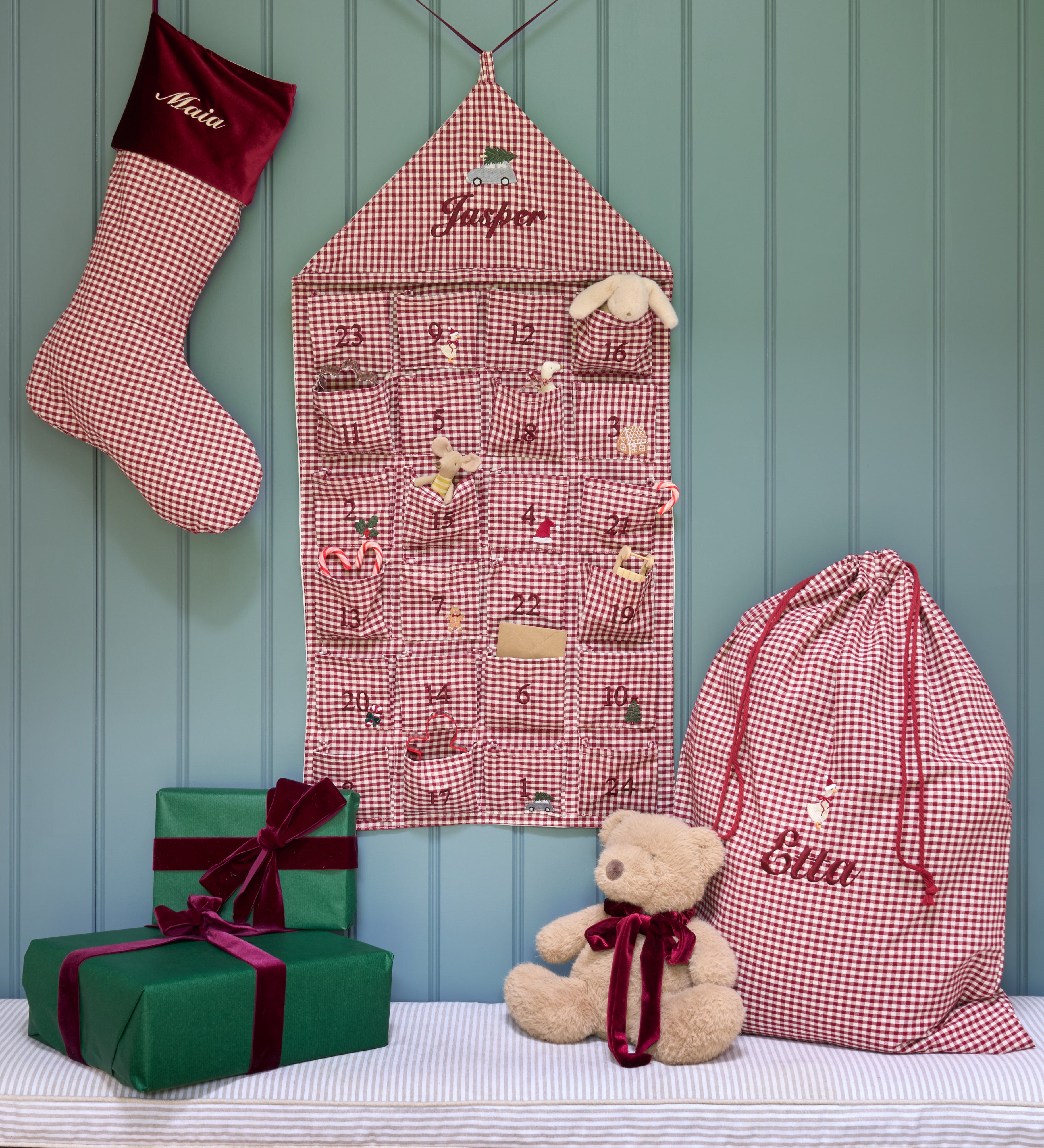 A festive Christmas advent setup featuring a burgundy and white gingham stocking, a matching house-shaped advent calendar, a large drawstring gift bag, and wrapped presents on a bench with a teddy bear, all set against a teal paneled wall.