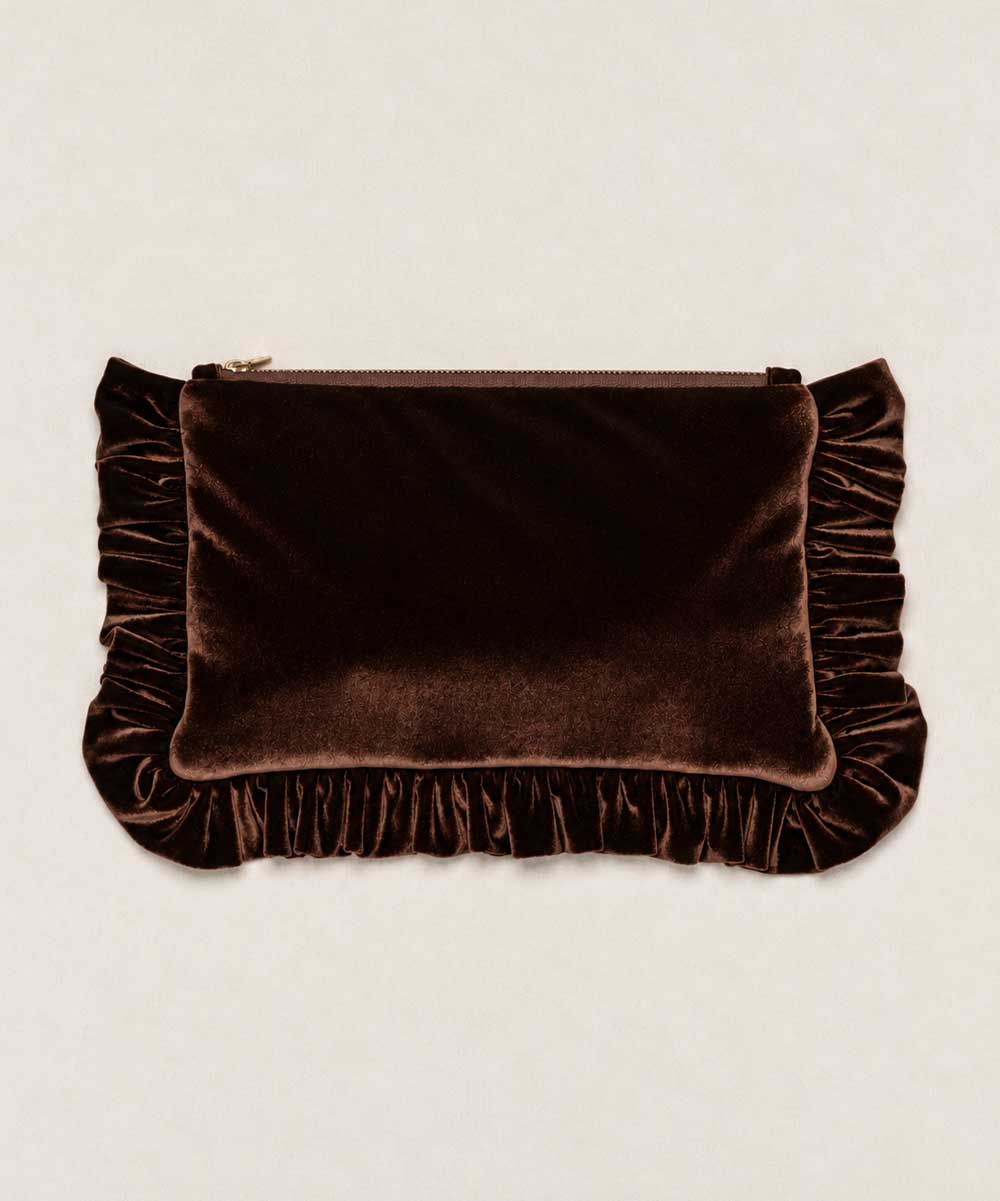 A dark brown velvet clutch bag with ruffled trim on the sides and bottom, featuring a top zipper closure, set against a light beige textured background.
