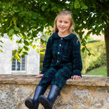 Young girl in black watch tartan pyjamas and rain boots sitting on a stone wall with greenery in the background
