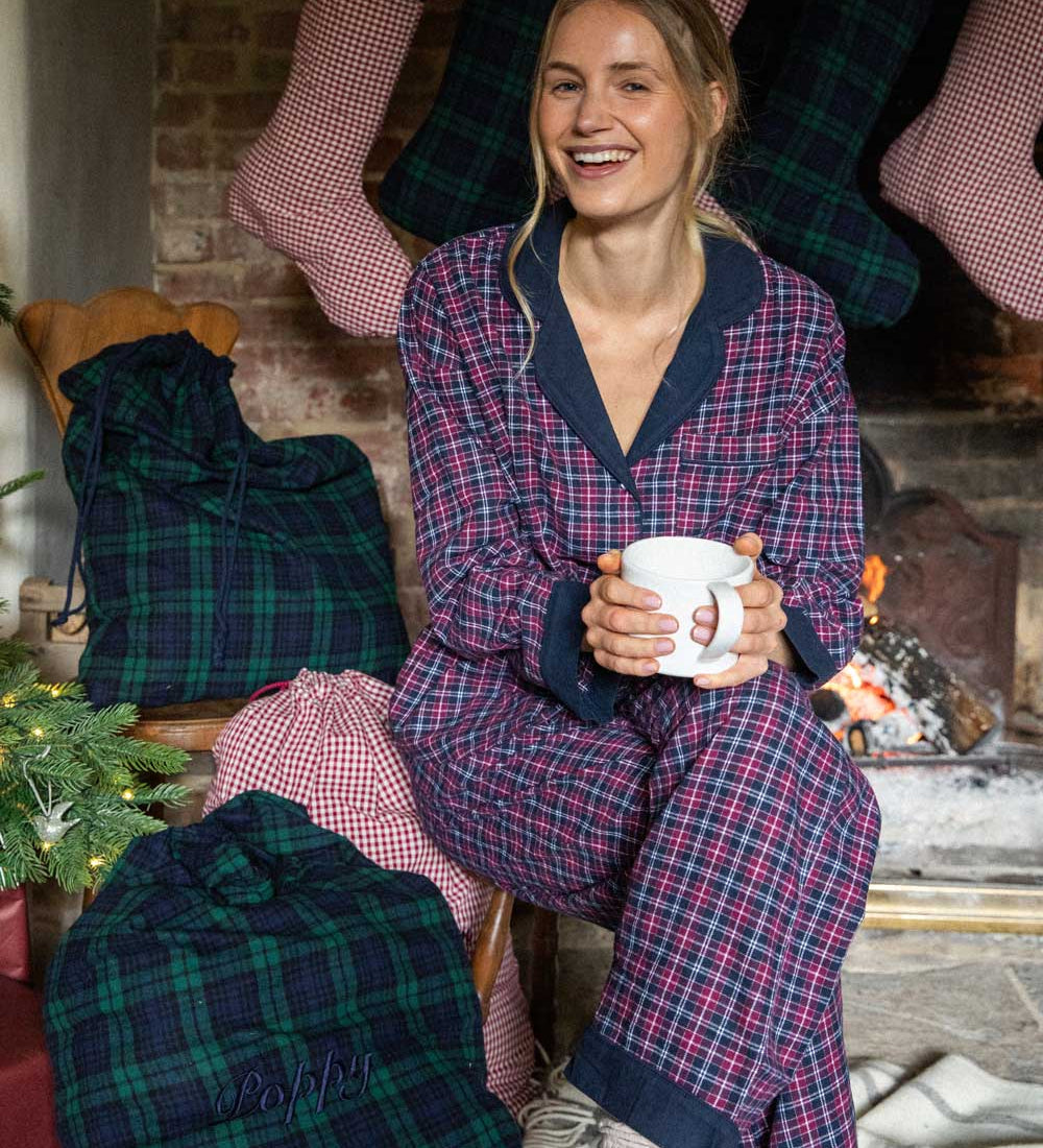 Woman in plaid pajamas holding a mug in a festive setting with stockings and a fireplace.