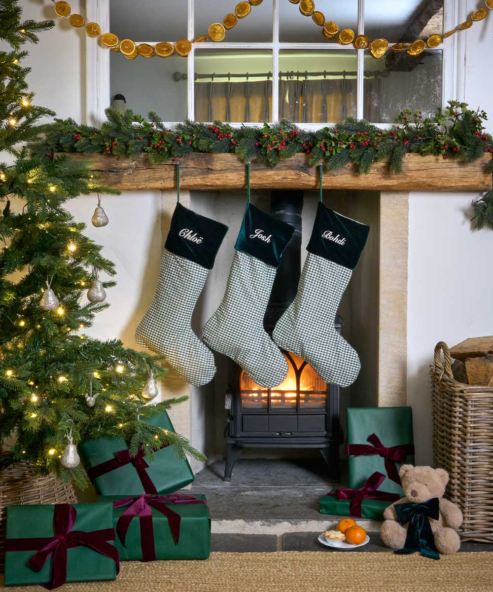 Three personalised Christmas stockings with green and white gingham patterns and forest green velvet cuffs hang from a wooden mantel decorated with a green garland. A lit fireplace is visible in the background, with a Christmas tree, wrapped presents, a teddy bear, and a wicker basket on the floor in a cozy holiday setting.