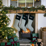 Three personalised Christmas stockings with green and white gingham patterns and forest green velvet cuffs hang from a wooden mantel decorated with a green garland. A lit fireplace is visible in the background, with a Christmas tree, wrapped presents, a teddy bear, and a wicker basket on the floor in a cozy holiday setting.