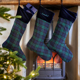 Three Christmas stockings with green and blue tartan plaid and dark navy velvet cuffs hang from a rustic wooden mantel above a burning wood-burning stove. One stocking is personalized with the name 'Gabriel' in white script.