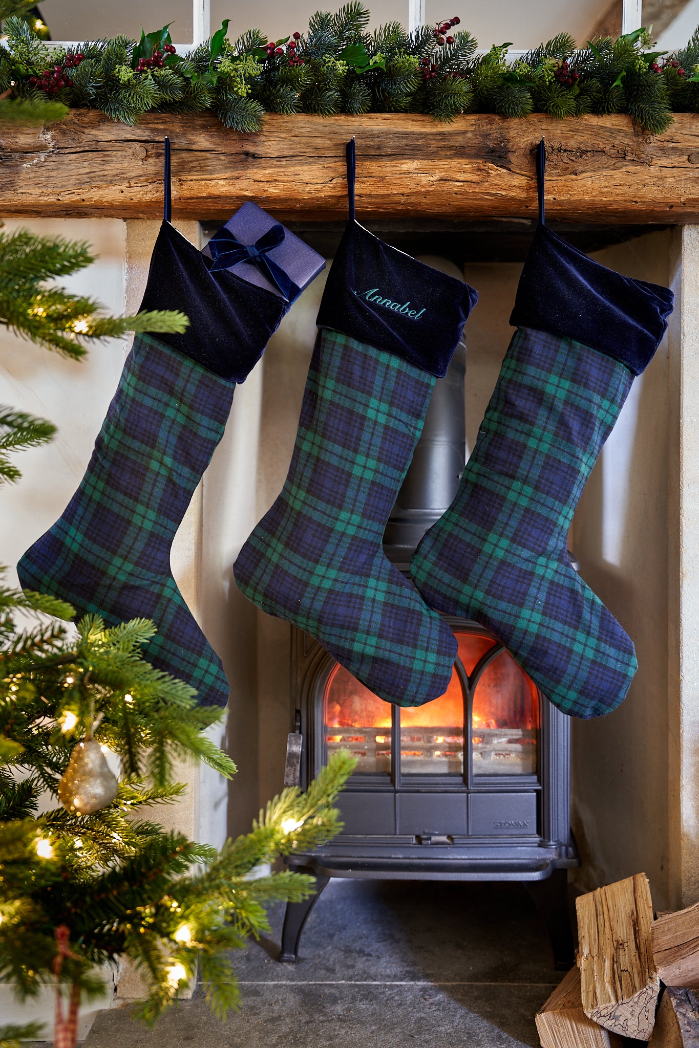 Three Christmas stockings with green and blue tartan plaid and dark navy velvet cuffs hang from a rustic wooden mantel above a burning wood-burning stove. One stocking is personalized with the name 'Gabriel' in white script.