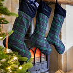 Three Christmas stockings with green and navy plaid fabric hang from a rustic wooden mantel above a black wood-burning stove. One stocking has a dark navy velvet cuff with a bow, and another has white embroidery.