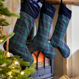 Three Christmas stockings with green and navy plaid fabric hang from a rustic wooden mantel above a black wood-burning stove. One stocking has a dark navy velvet cuff with a bow, and another has white embroidery.