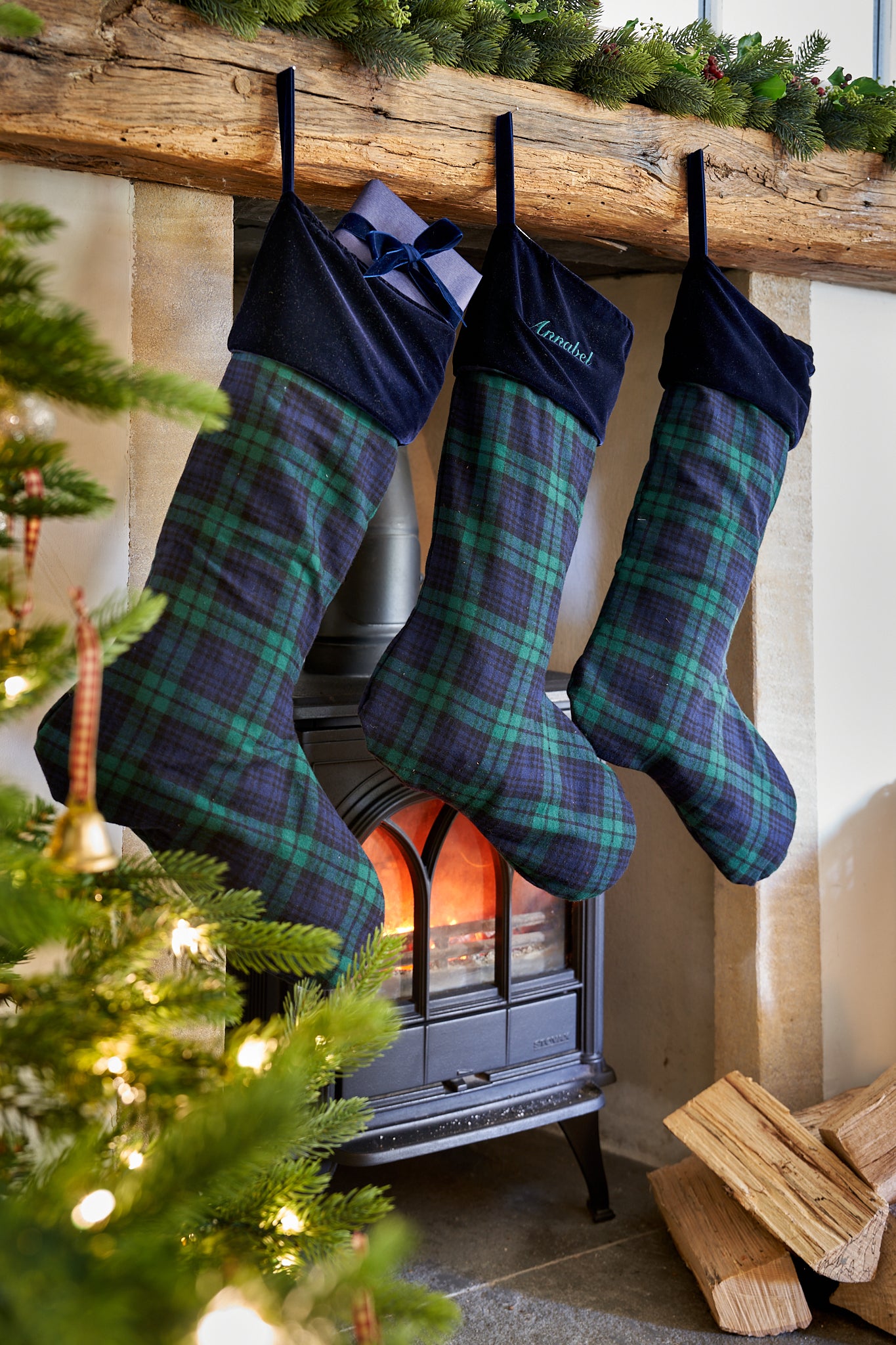 Three Christmas stockings with green and navy plaid fabric hang from a rustic wooden mantel above a black wood-burning stove. One stocking has a dark navy velvet cuff with a bow, and another has white embroidery.
