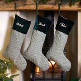 Three personalized Christmas stockings with green velvet cuffs and green-and-white gingham bodies hang from a wooden mantel above a burning fireplace. The names 'Bebidi', 'Josh', and 'Ghose' are embroidered in white on the cuffs.