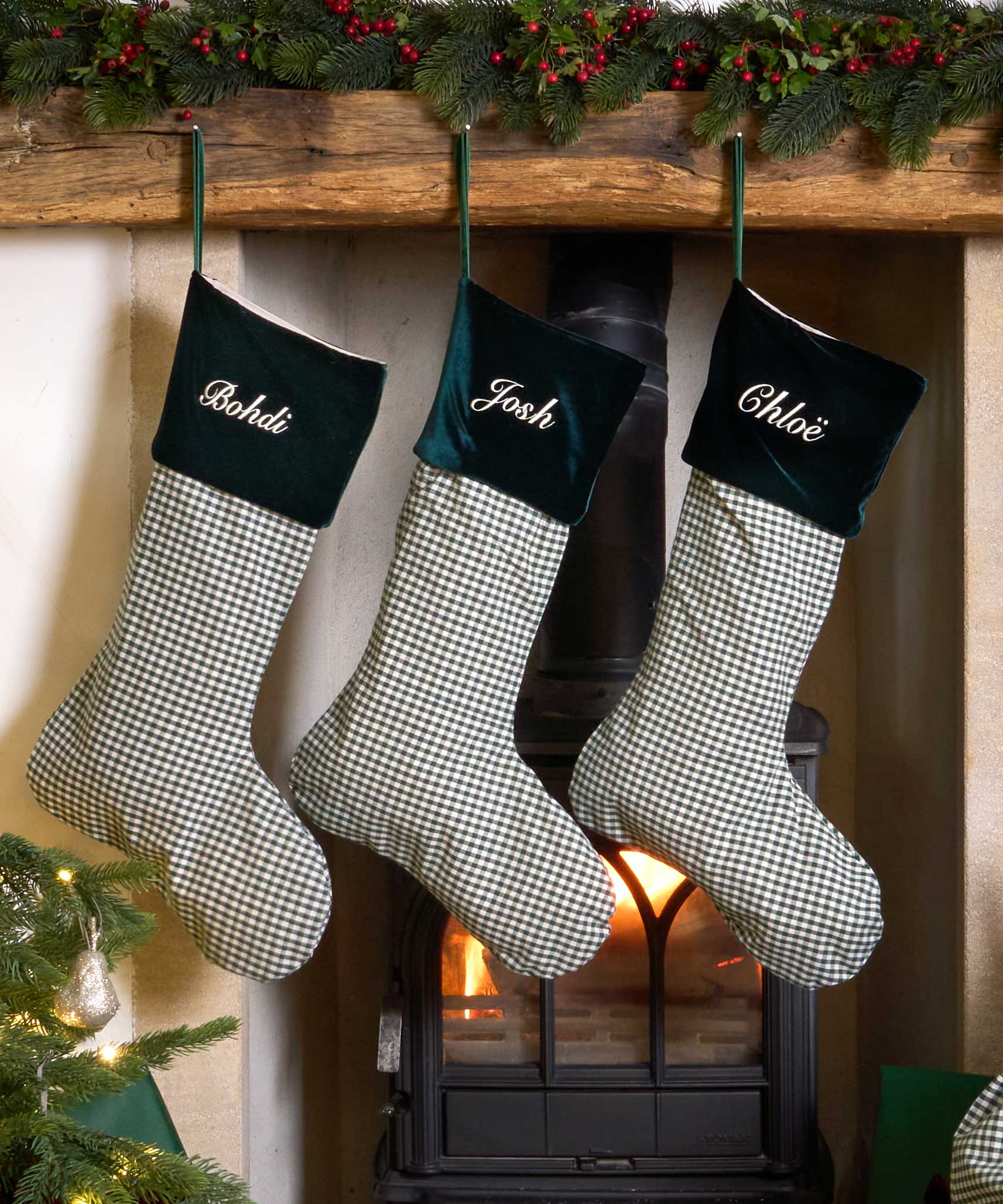 Three personalized Christmas stockings with green velvet cuffs and green-and-white gingham bodies hang from a wooden mantel above a burning fireplace. The names 'Bebidi', 'Josh', and 'Ghose' are embroidered in white on the cuffs.