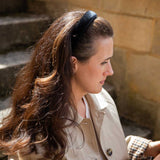 A woman with long brown hair wearing a black fabric headband, styled in a side-profile portrait against a warm stone wall background.