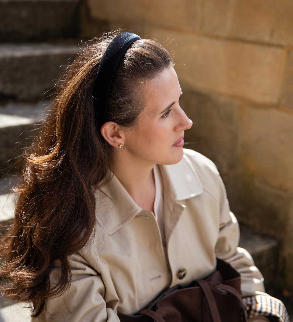 A woman with long brown hair wearing a black headband, a beige trench coat, and holding a dark brown bag, sitting outdoors against a stone wall.