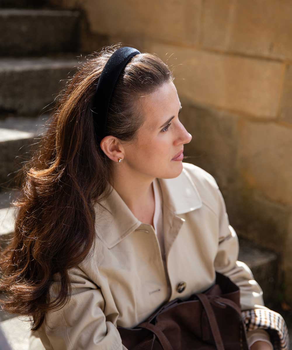 A woman with long brown hair wearing a black headband, a beige trench coat, and holding a dark brown bag, sitting outdoors against a stone wall.
