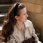 A woman with long brown hair wearing a black headband, a beige trench coat, and holding a dark brown bag, sitting outdoors against a stone wall.
