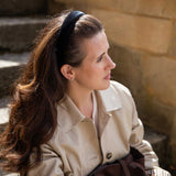 A woman with long brown hair wearing a black headband, a beige trench coat, and holding a dark brown bag, sitting outdoors against a stone wall.