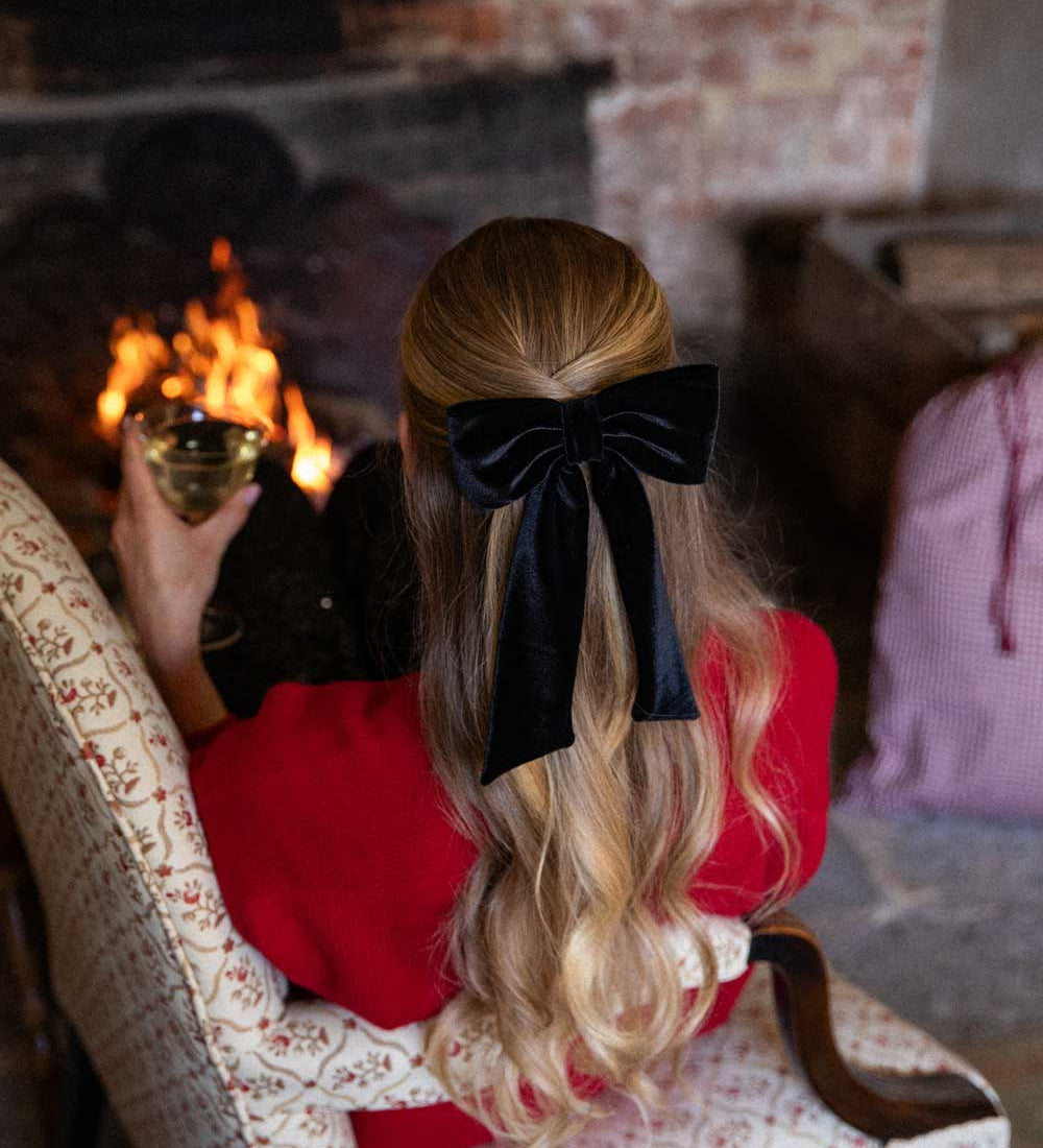 Person with a black velvet hair bow in their hair sitting by a fireplace holding a glass of wine.