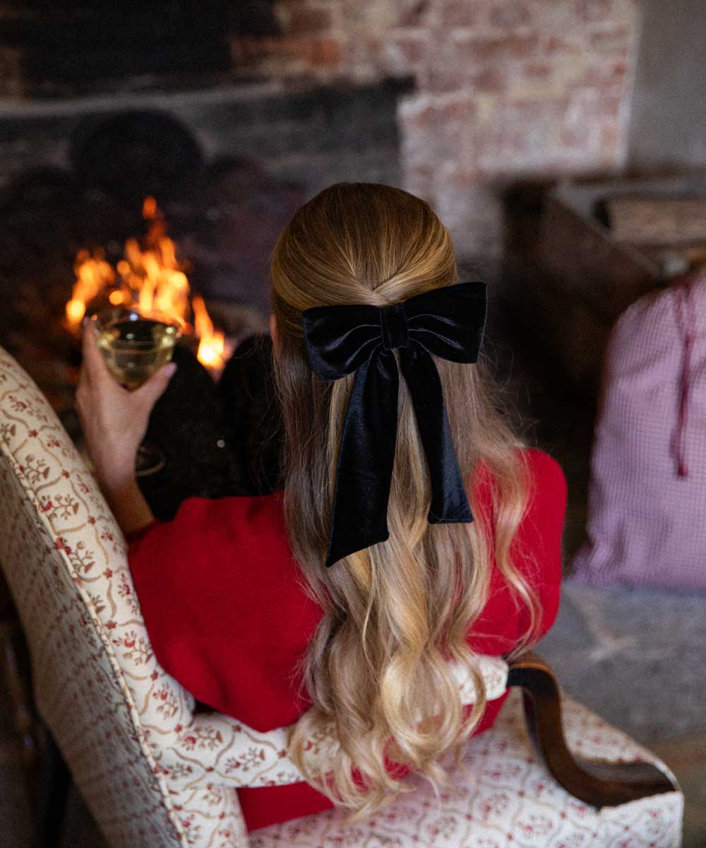 Person with a black velvet hair bow in their hair sitting by a fireplace holding a glass of wine.