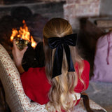Person with a black velvet hair bow in their hair sitting by a fireplace holding a glass of wine.