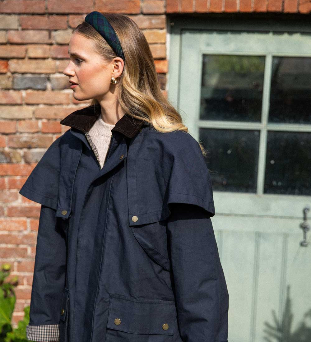 Woman wearing a navy blue coat standing in front of a brick wall and glass door wearing a tartan headband