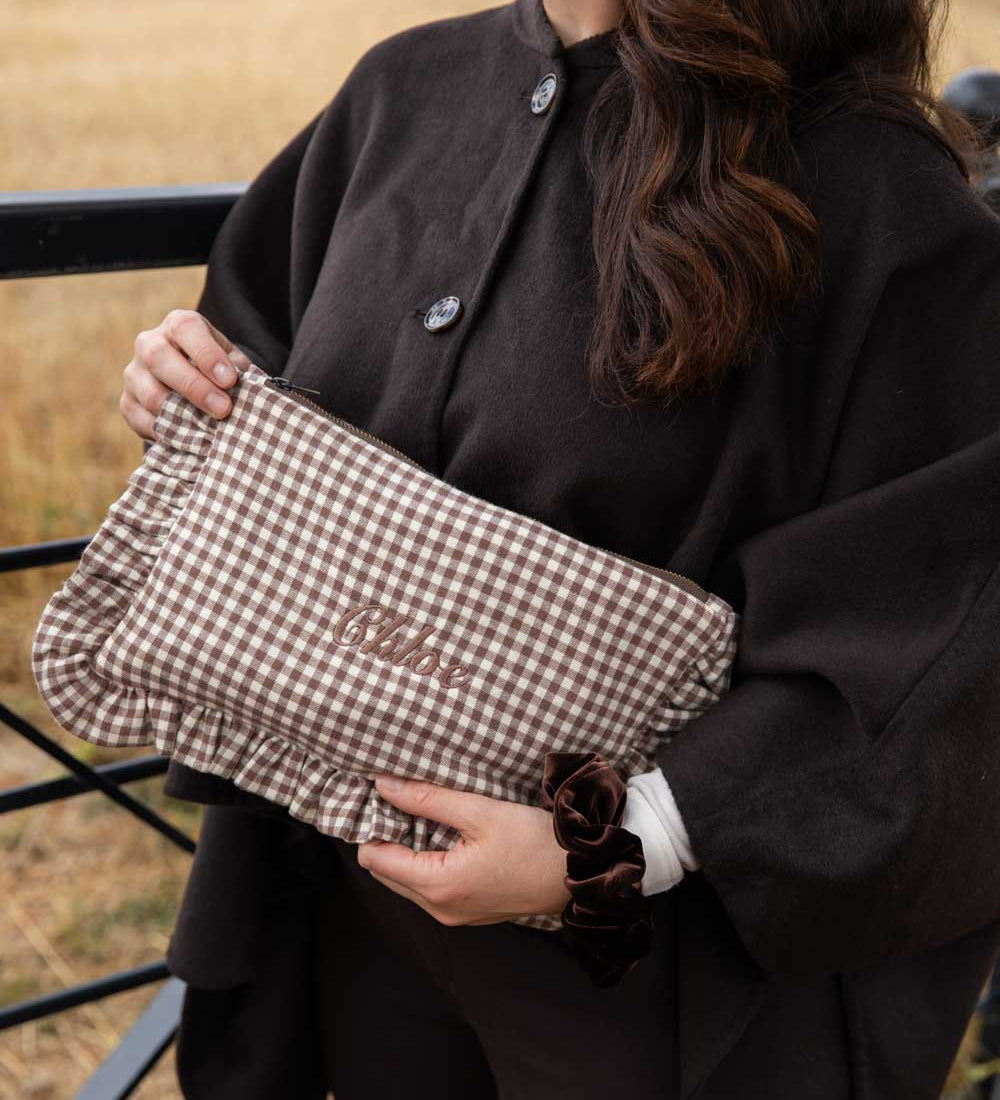 A person outdoors holding a brown and beige gingham clutch bag with the name 'chloe' embroidered on the front. The person is wearing a dark brown coat.