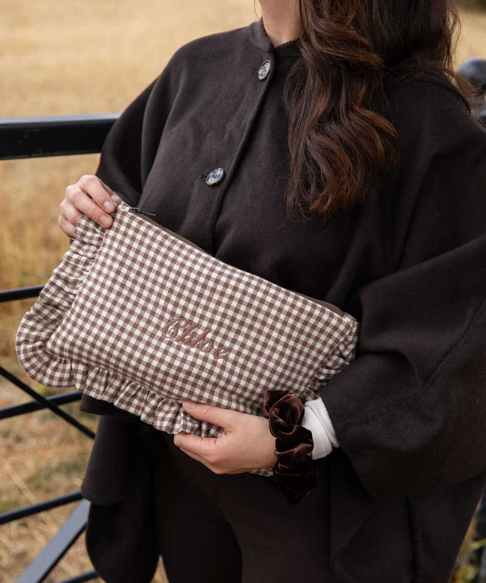 A person outdoors holding a brown and beige gingham clutch bag with the name 'chloe' embroidered on the front. The person is wearing a dark brown coat.