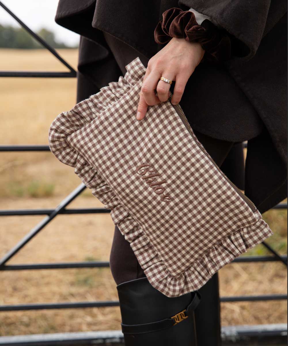 A person outdoors holds a brown and beige gingham clutch with ruffled edges and the word 'chloe embroidered on the front. The person is wearing a dark coat, black leggings, and black rain boots, with a dry field and metal gate in the background.